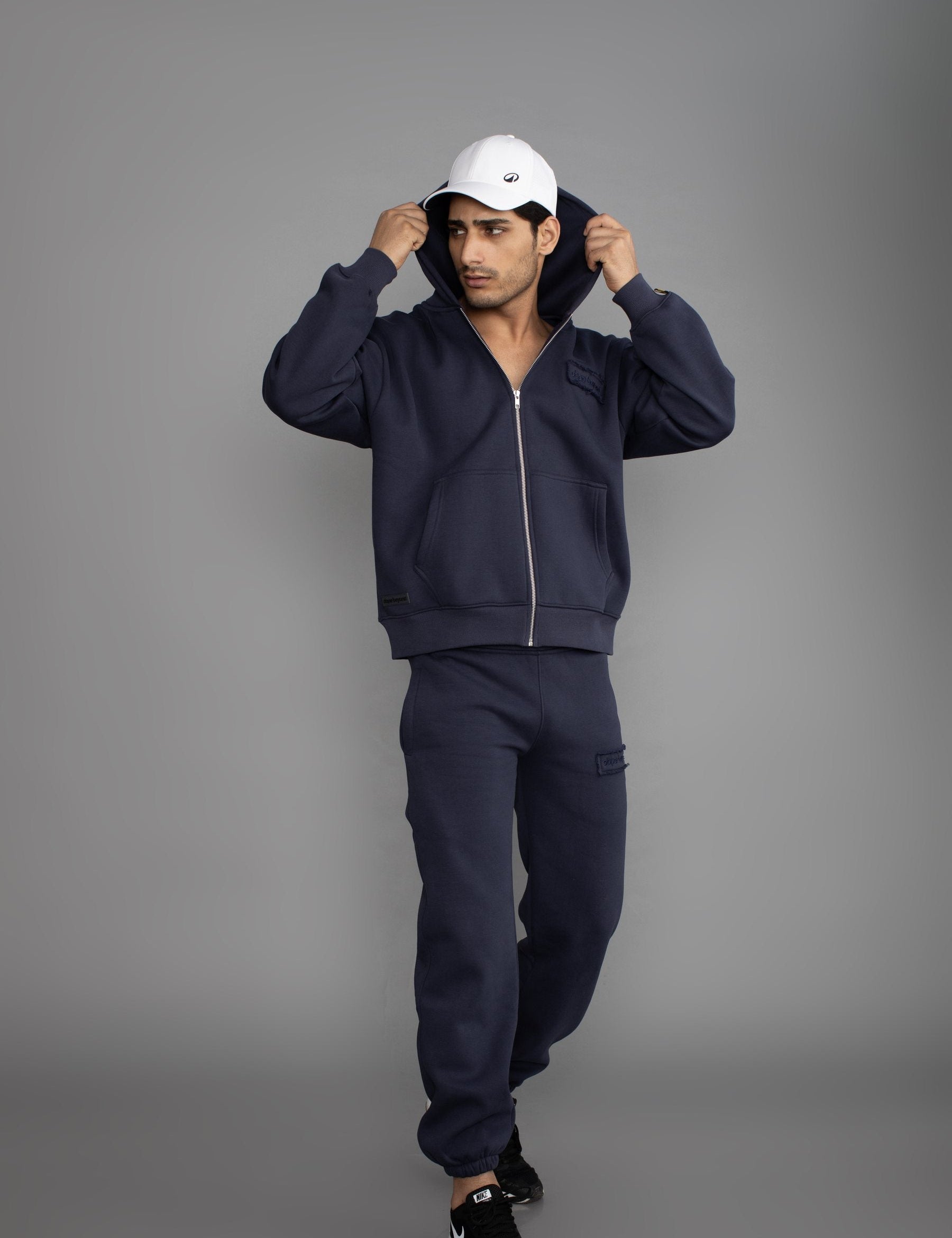 Man in a white cap and the Dope Beyond Ink Navy zip hoodie and joggers set, full length, studio