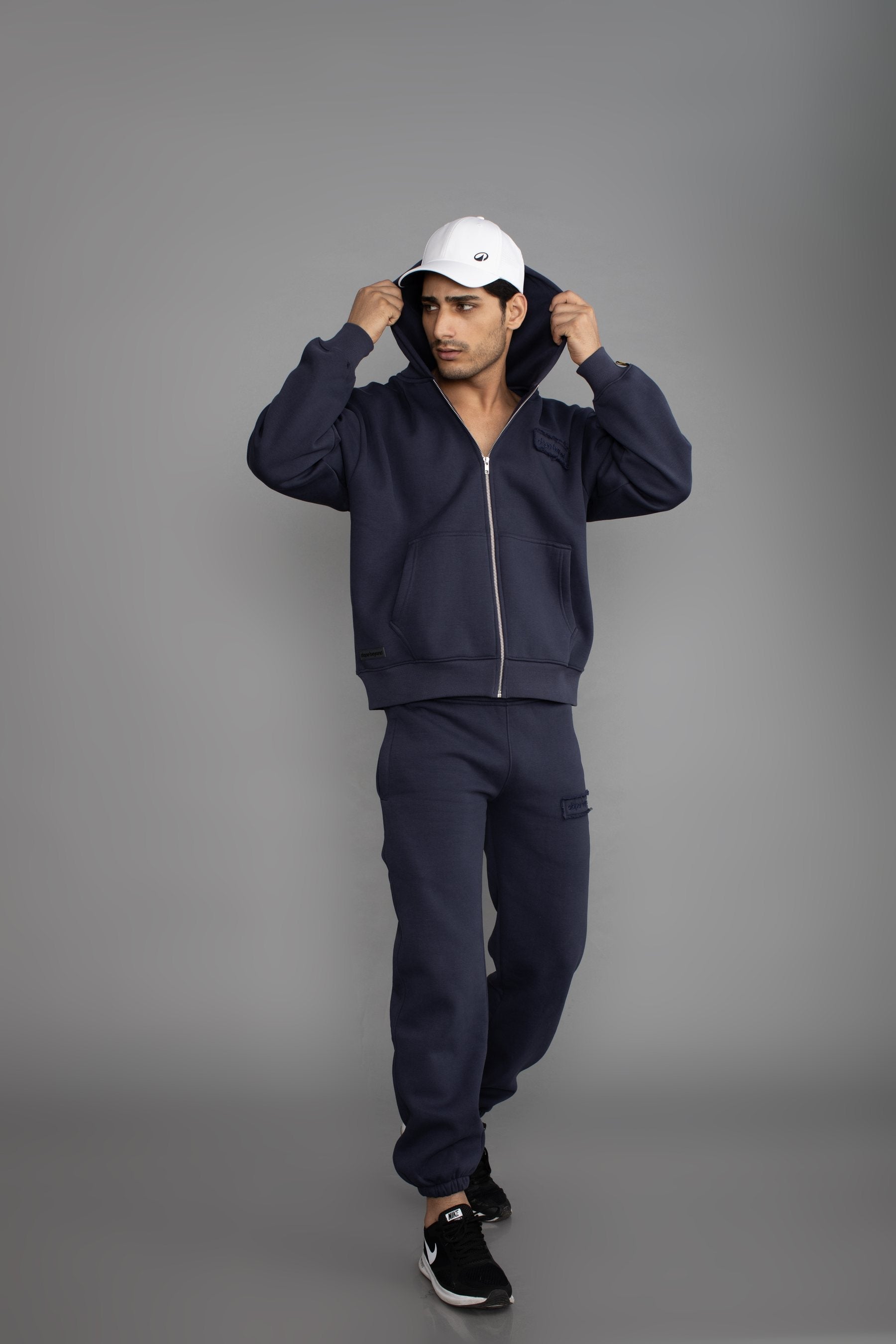 Man in a white cap and the Dope Beyond Ink Navy zip hoodie and joggers set, full length, studio