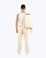 Man in beige heavyweight joggers and white tank, holding 'beyond' logo sweatshirt, dopebeyond streetwear