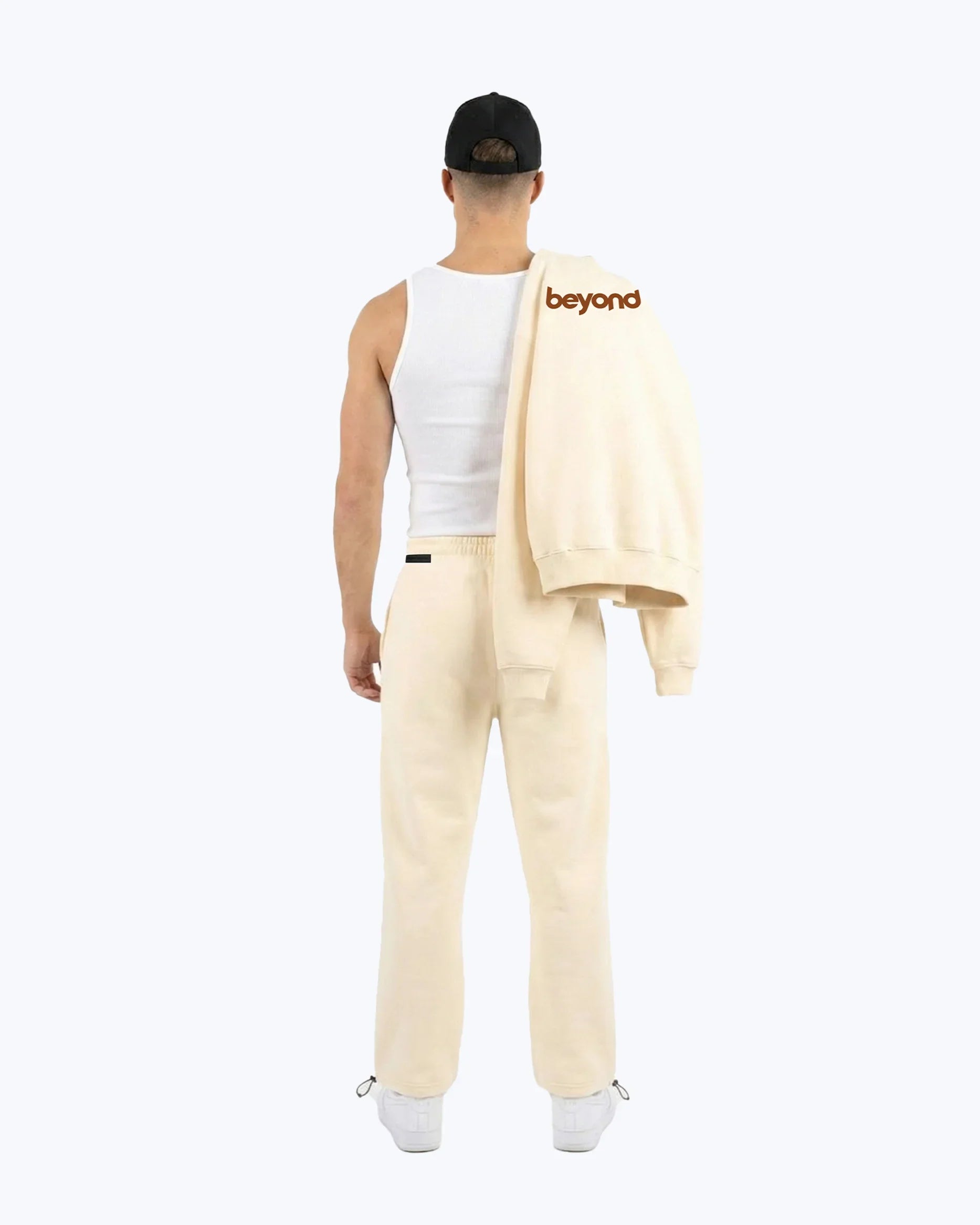 Man in beige heavyweight joggers and white tank, holding 'beyond' logo sweatshirt, dopebeyond streetwear