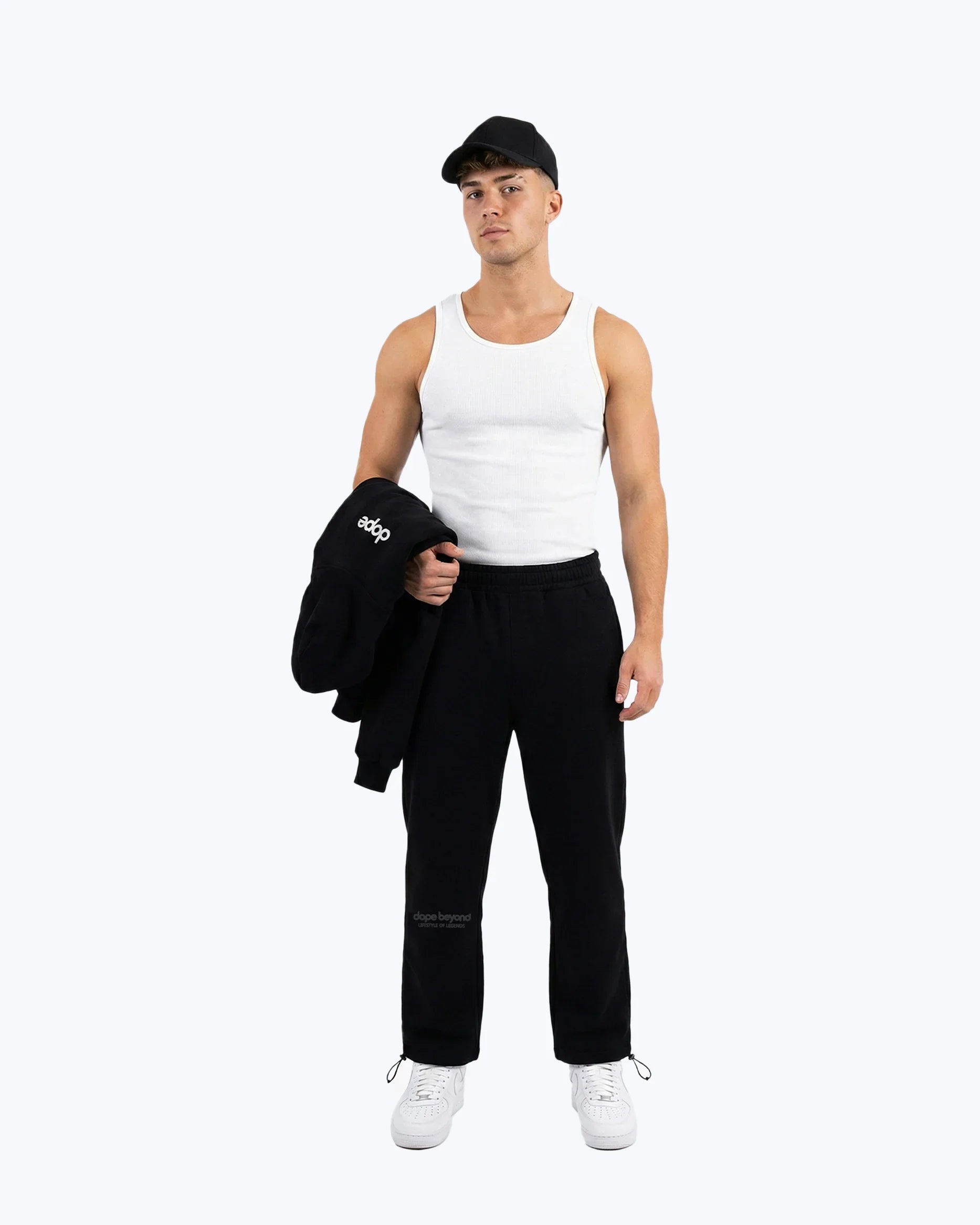 Man wearing Dope Beyond black heavyweight joggers, white tank top, black cap, and holding hoodie