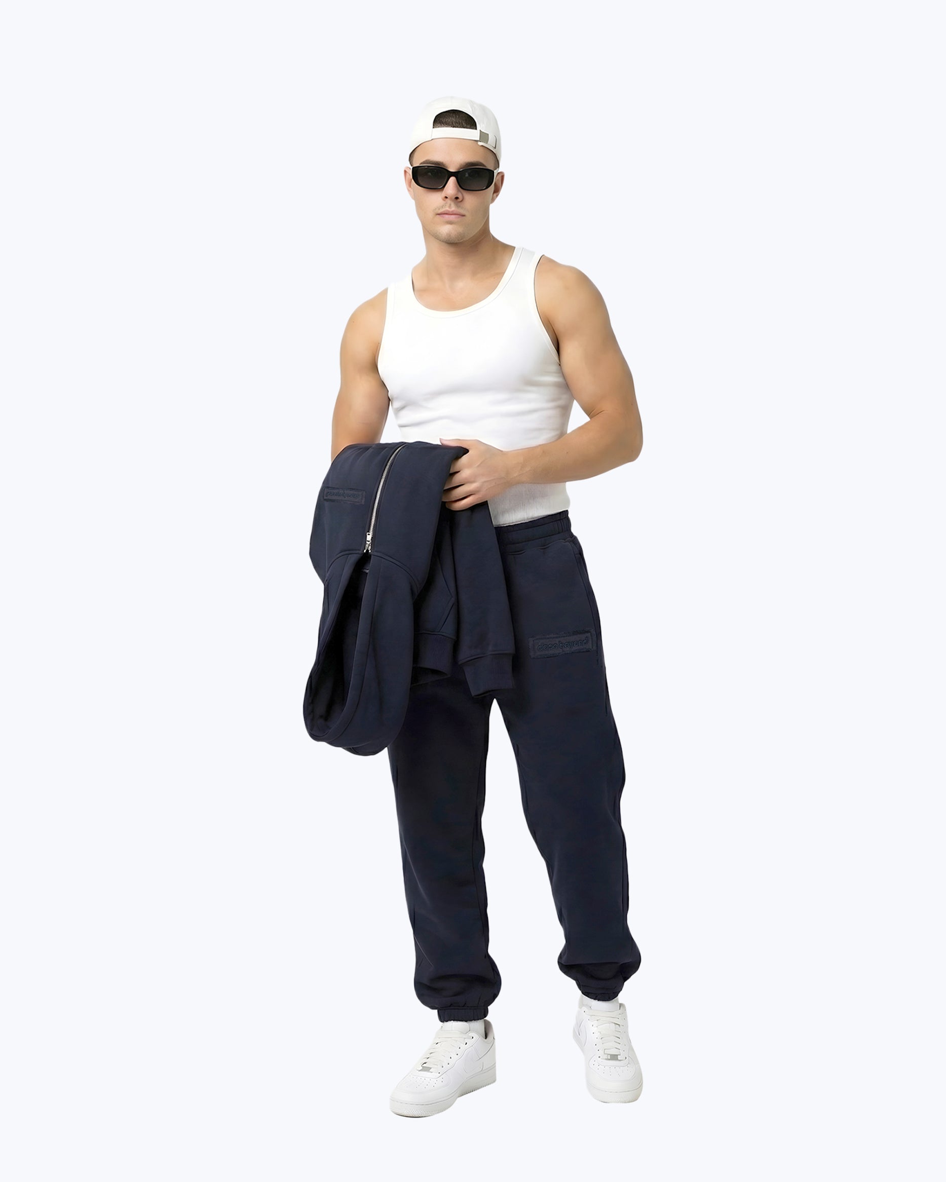 Man wearing Dope Beyond Ink navy heavyweight joggers, white tank top, cap, and sunglasses.