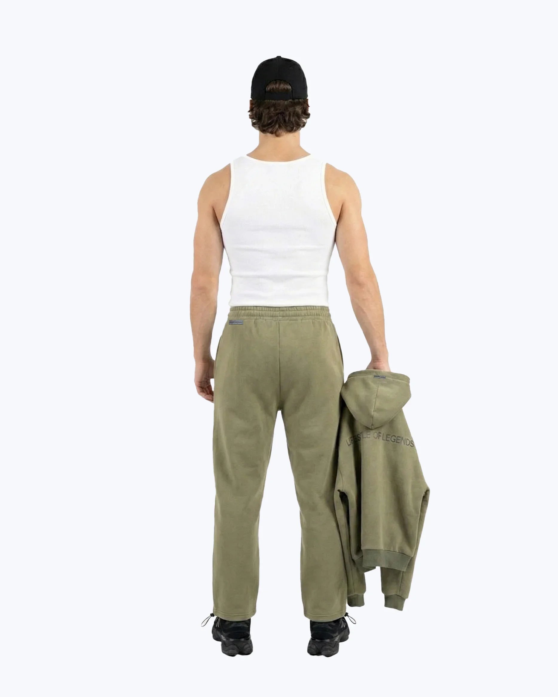 Man wearing olive green Dope Beyond gym joggers, white tank top, and black cap, holding matching hoodie