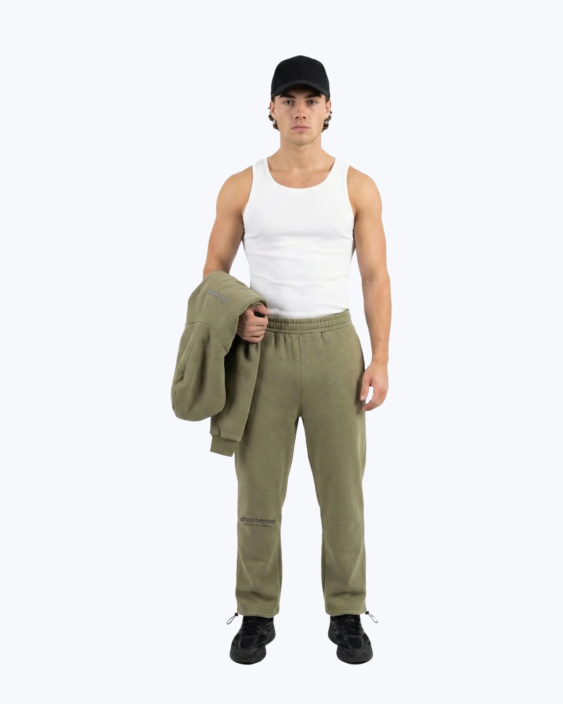 Man in olive green Dope Beyond gym joggers, white tank top, and black cap standing against plain background