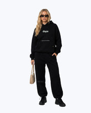 Woman wearing Dope Beyond black streetwear hoodie and joggers set, holding beige bag