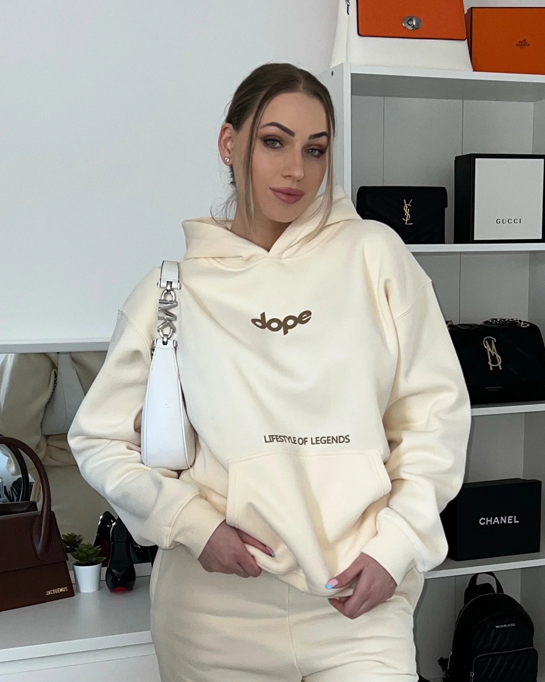 Woman wearing cream Dope Beyond hoodie with 'Lifestyle of Legends' text, luxury bags in background