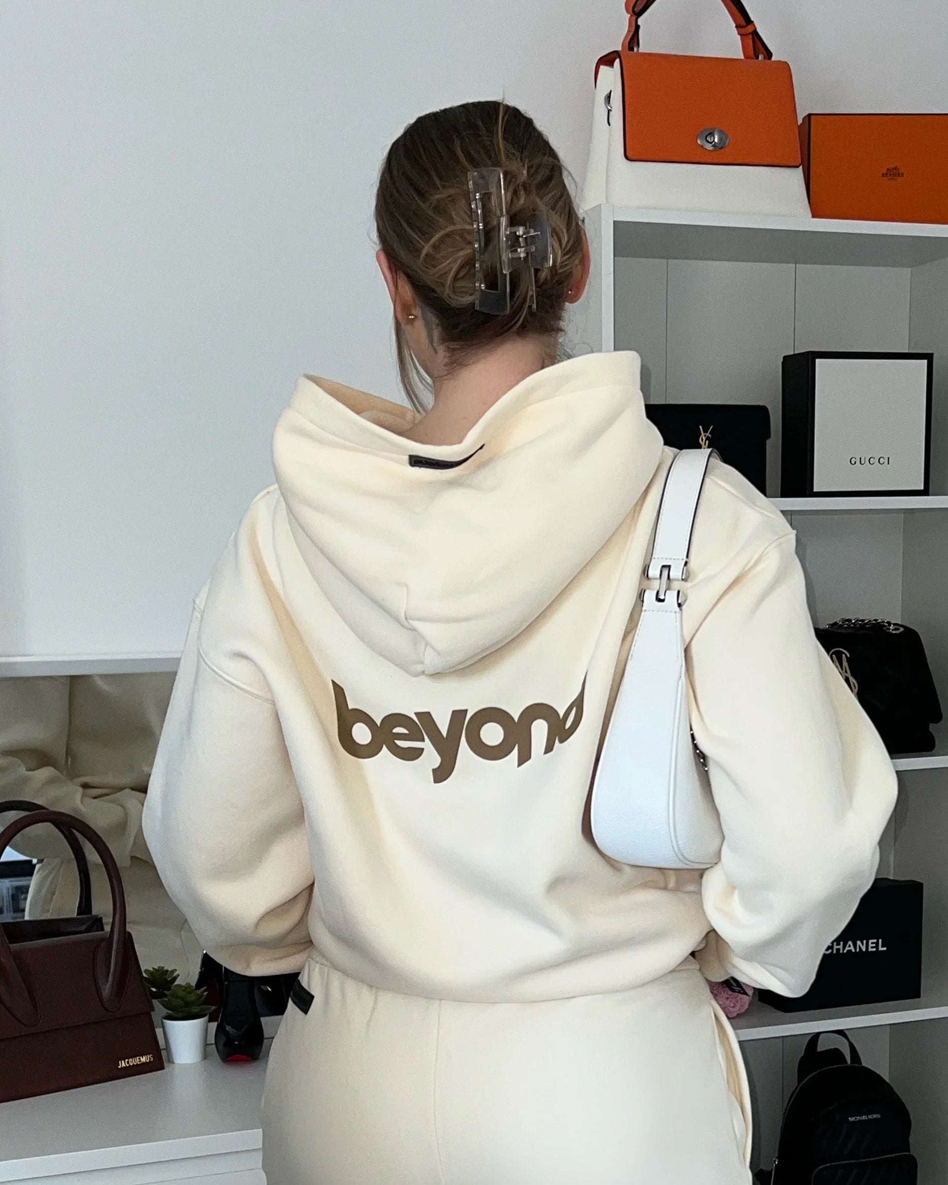 Woman in cream dopebeyond hoodie with 'beyond' logo, white purse, luxury bags in background