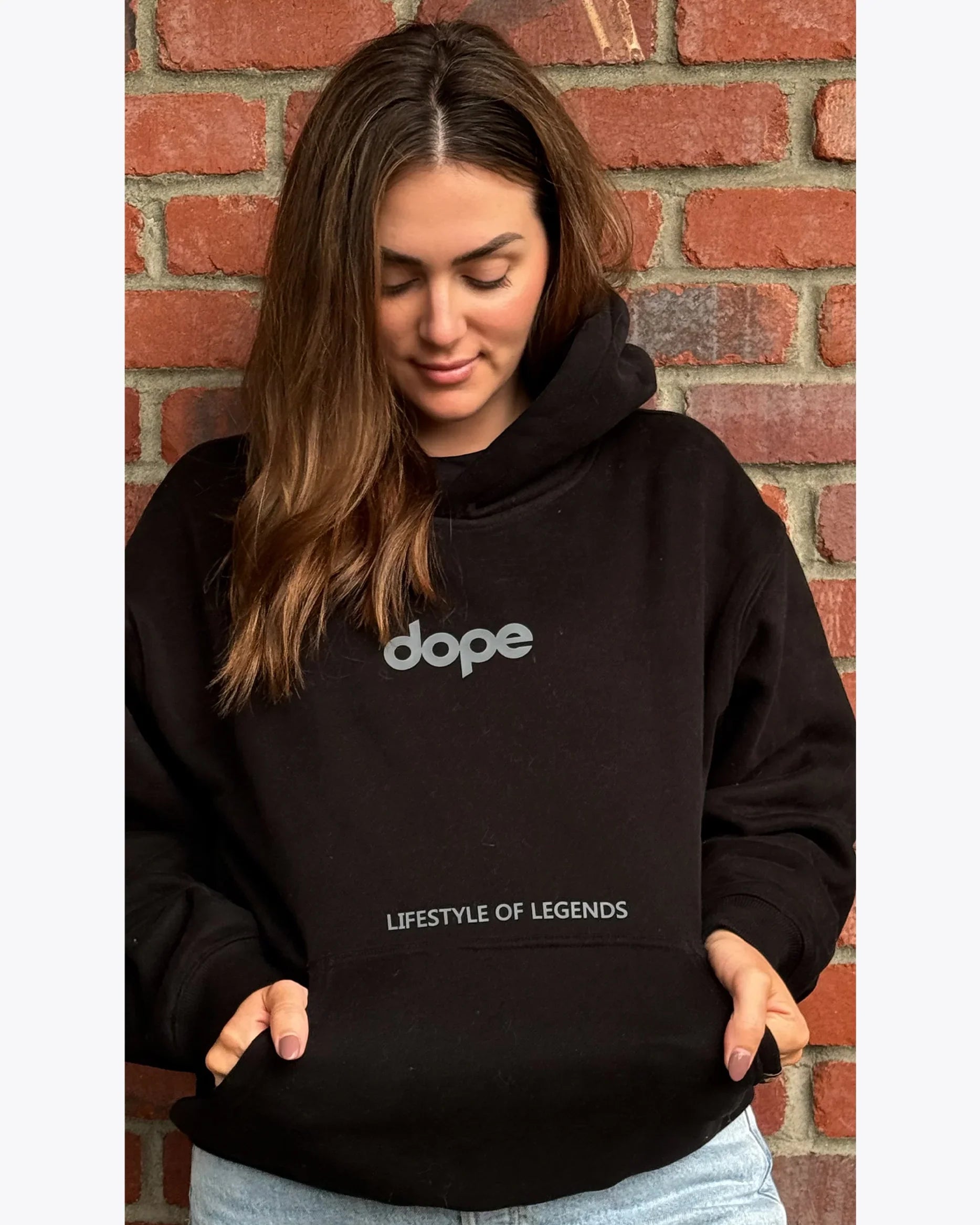 Woman wearing black dopebeyond streetwear hoodie with 'LIFESTYLE OF LEGENDS' text, brick wall background.